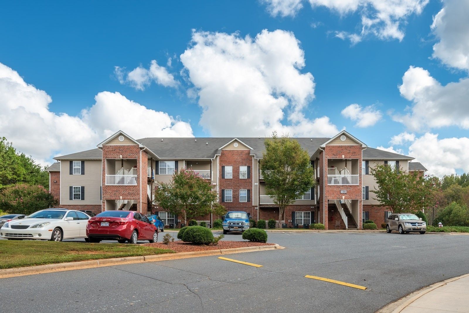 Apartment building exterior, beige with brick accents, cars parked out front, blue sky with clouds.