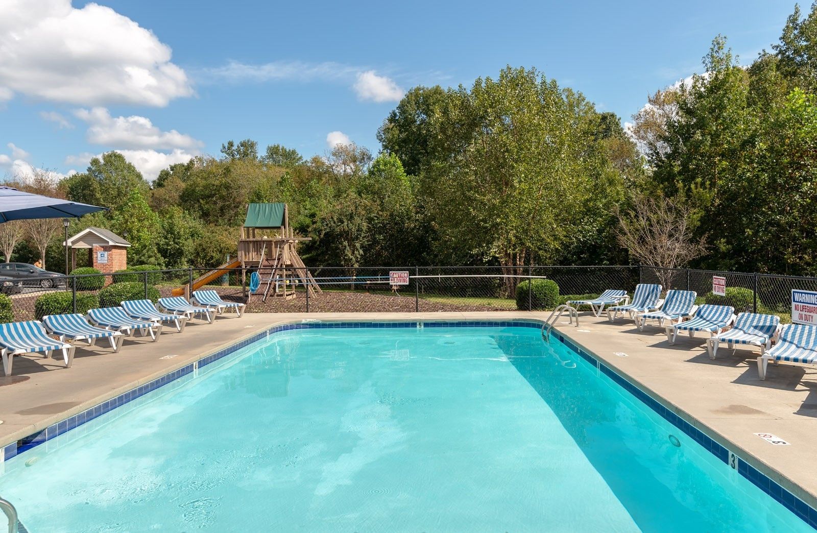 Swimming pool with blue and white striped lounge chairs, playground, and trees under a sunny sky.
