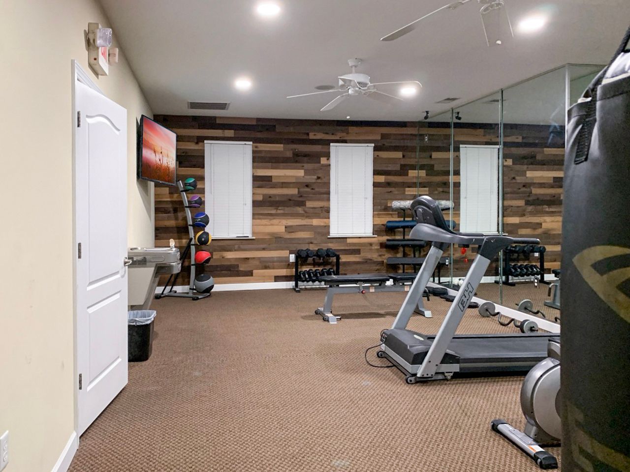 Home gym with exercise equipment: treadmill, weights, punching bag, wood accent wall, and a TV.