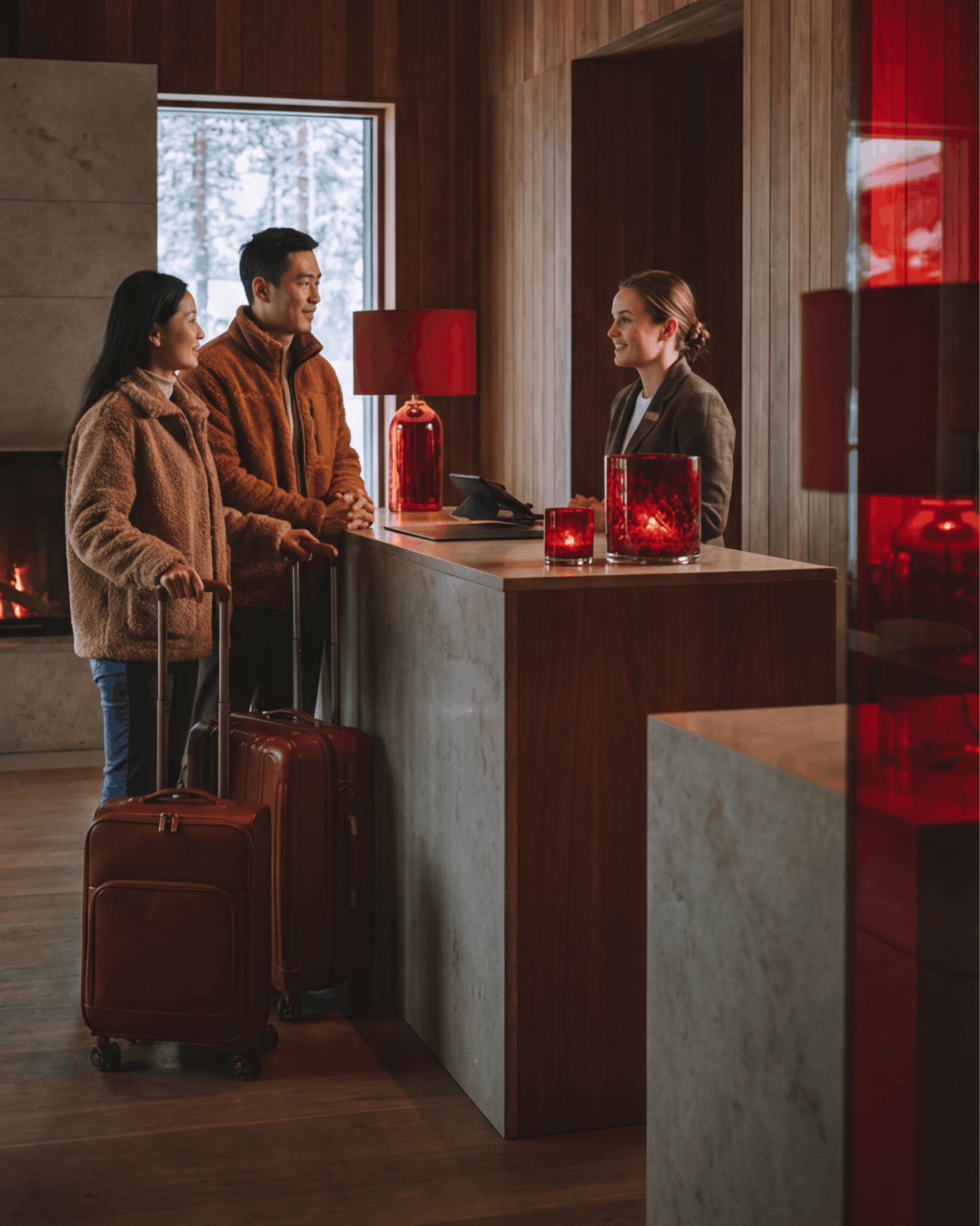 To personer med bagasje står ved en hotellresepsjon og snakker med en ansatt i en varm lobby med trepanel.