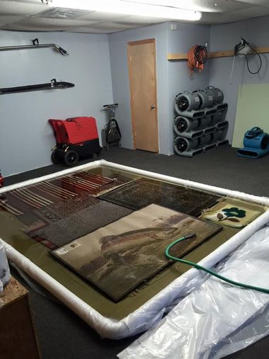 Flood-damaged garage with wet carpet, drying fan, dehumidifier, and equipment stacked against the wall