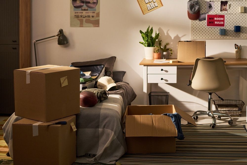 Cluttered bedroom with moving boxes, unmade bed, desk, chair, and plants in warm light