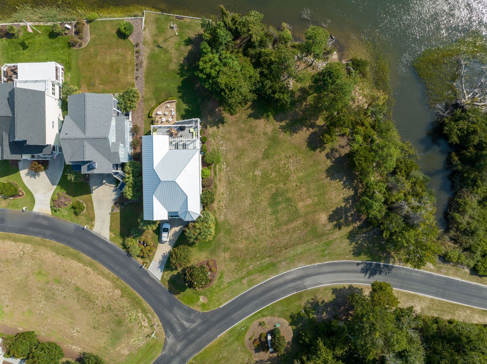 Aerial view: houses with driveways, vacant lot, curving road, and water with trees.