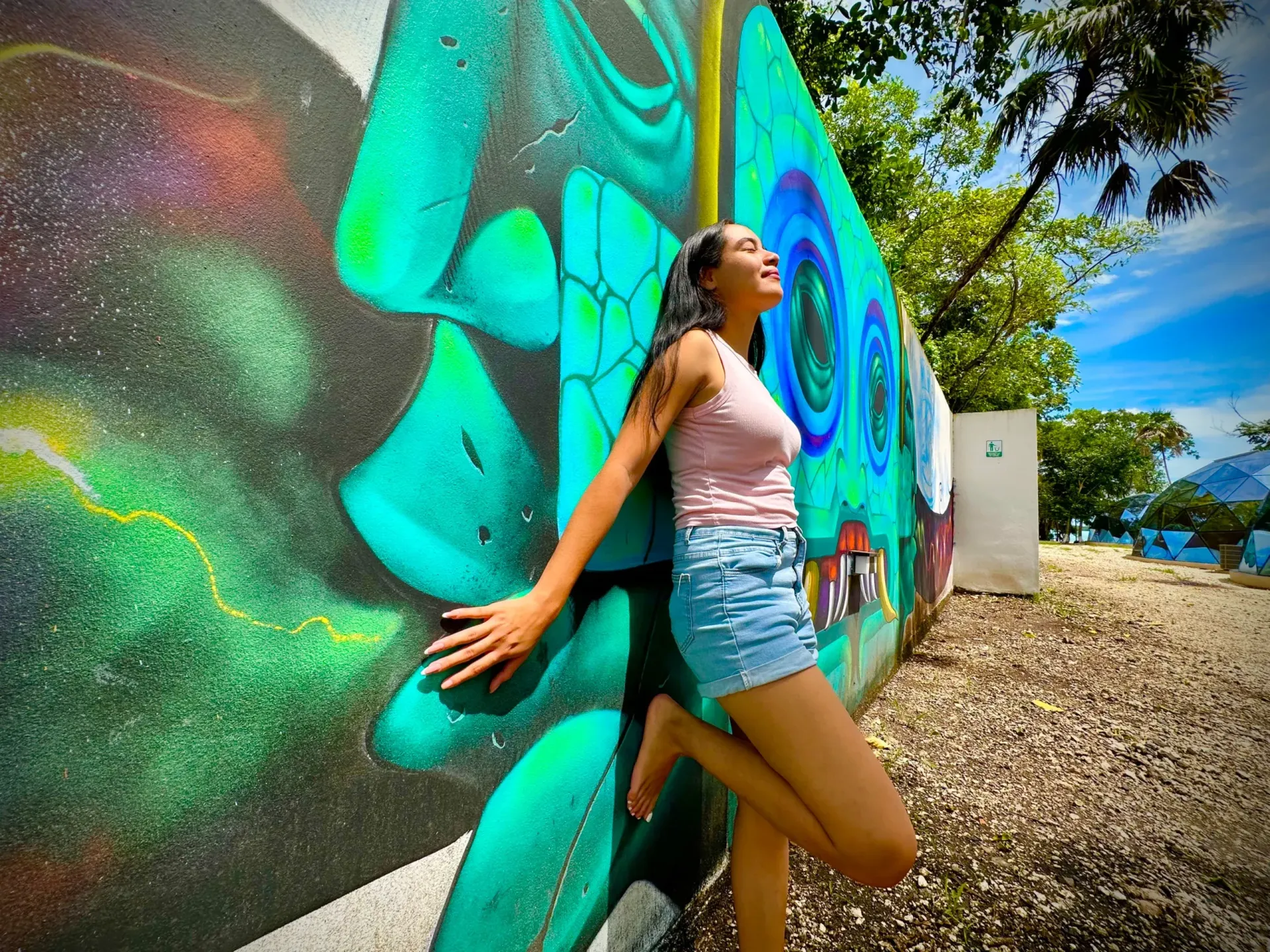 Una mujer se apoya en una pared con grafitis de colores, al aire libre. Lleva una blusa rosa, pantalones cortos vaqueros