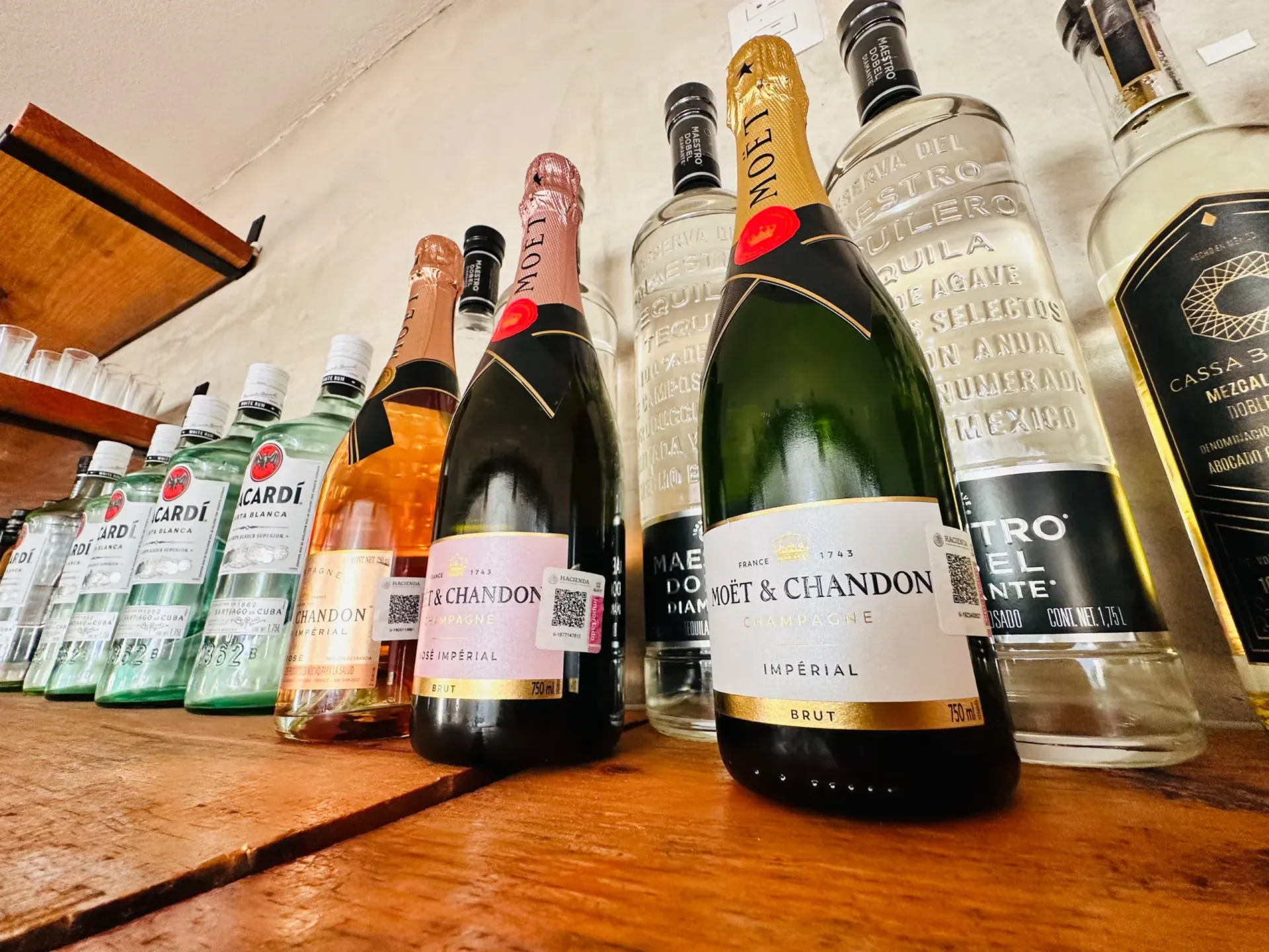 Botellas de alcohol expuestas en un estante de madera, incluyendo champán y tequila, con un montaje de bar.