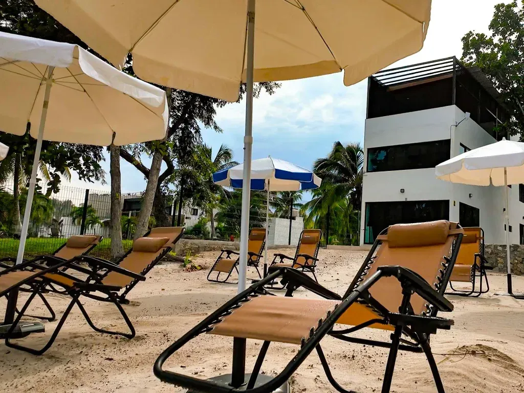Escena de playa con tumbonas bajo sombrillas, cerca de un edificio blanco con ventanas oscuras.