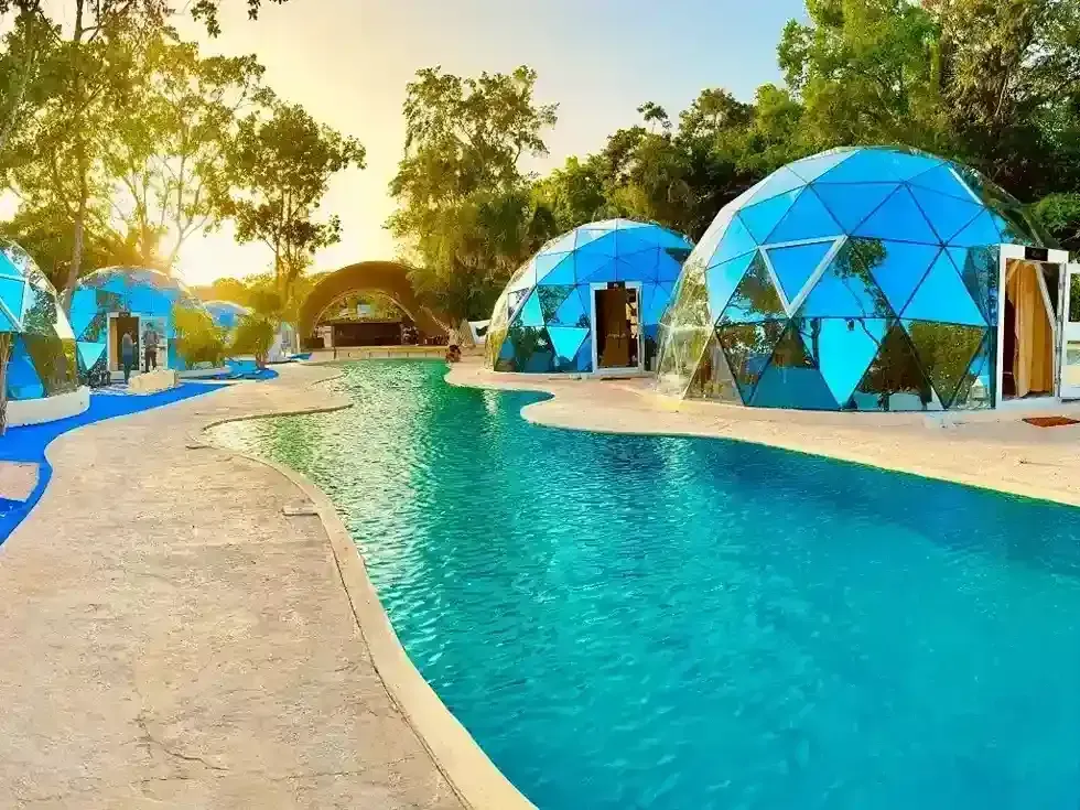 Estructuras de cúpulas geodésicas azules junto a una piscina turquesa, en un entorno soleado al aire libre.