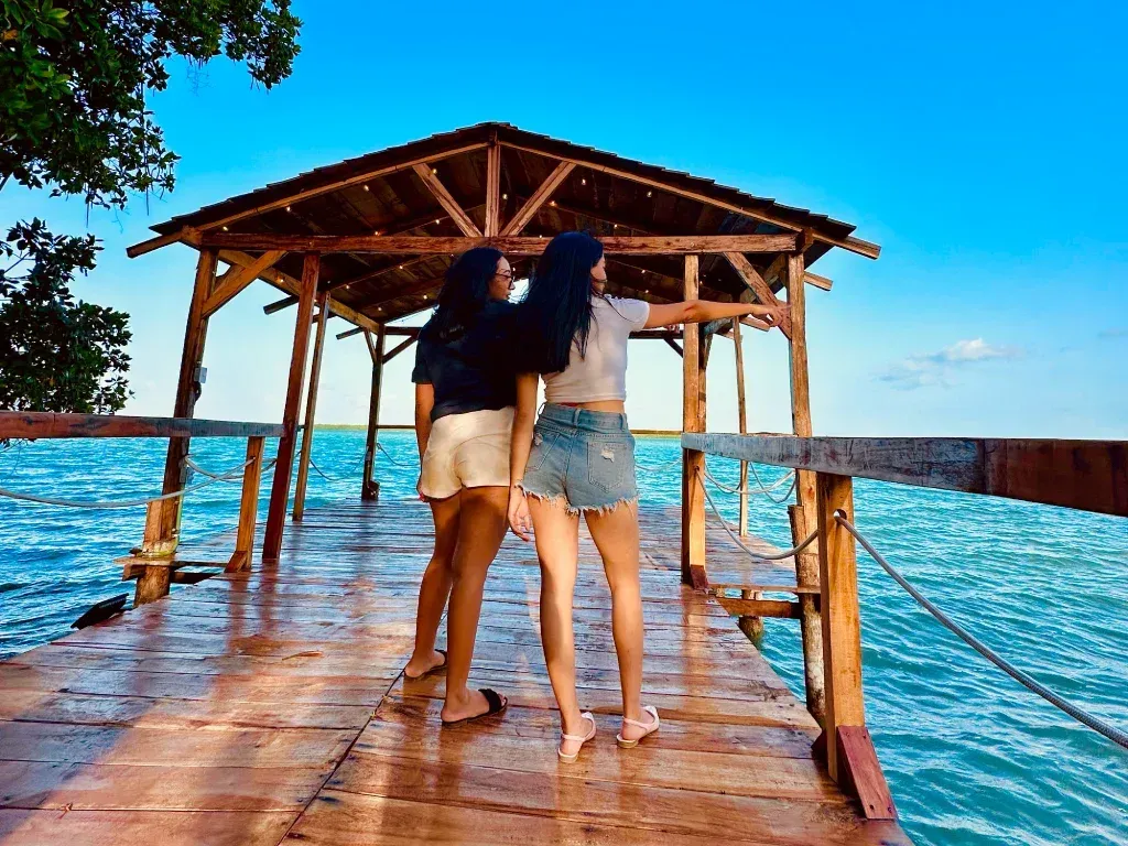 Dos mujeres de pie sobre un muelle de madera sobre aguas turquesas, señalando hacia el horizonte en un techo de madera.