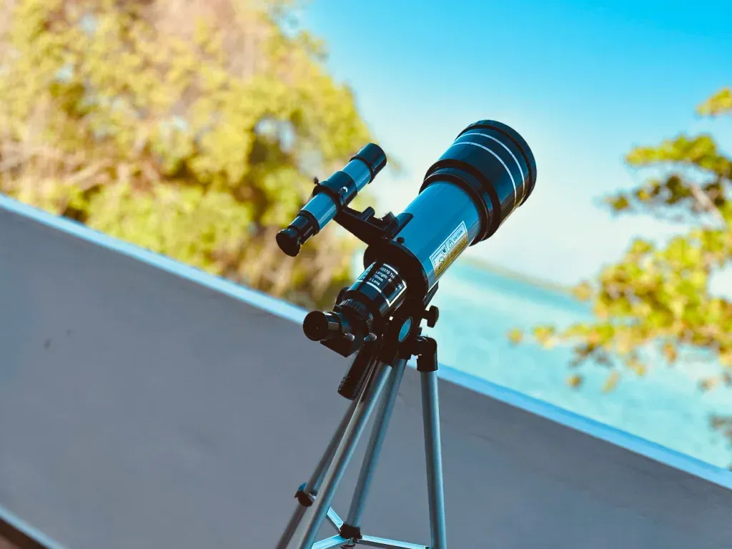 Telescopio sobre trípode, apuntando hacia el cielo con un fondo oceánico azul y verde.