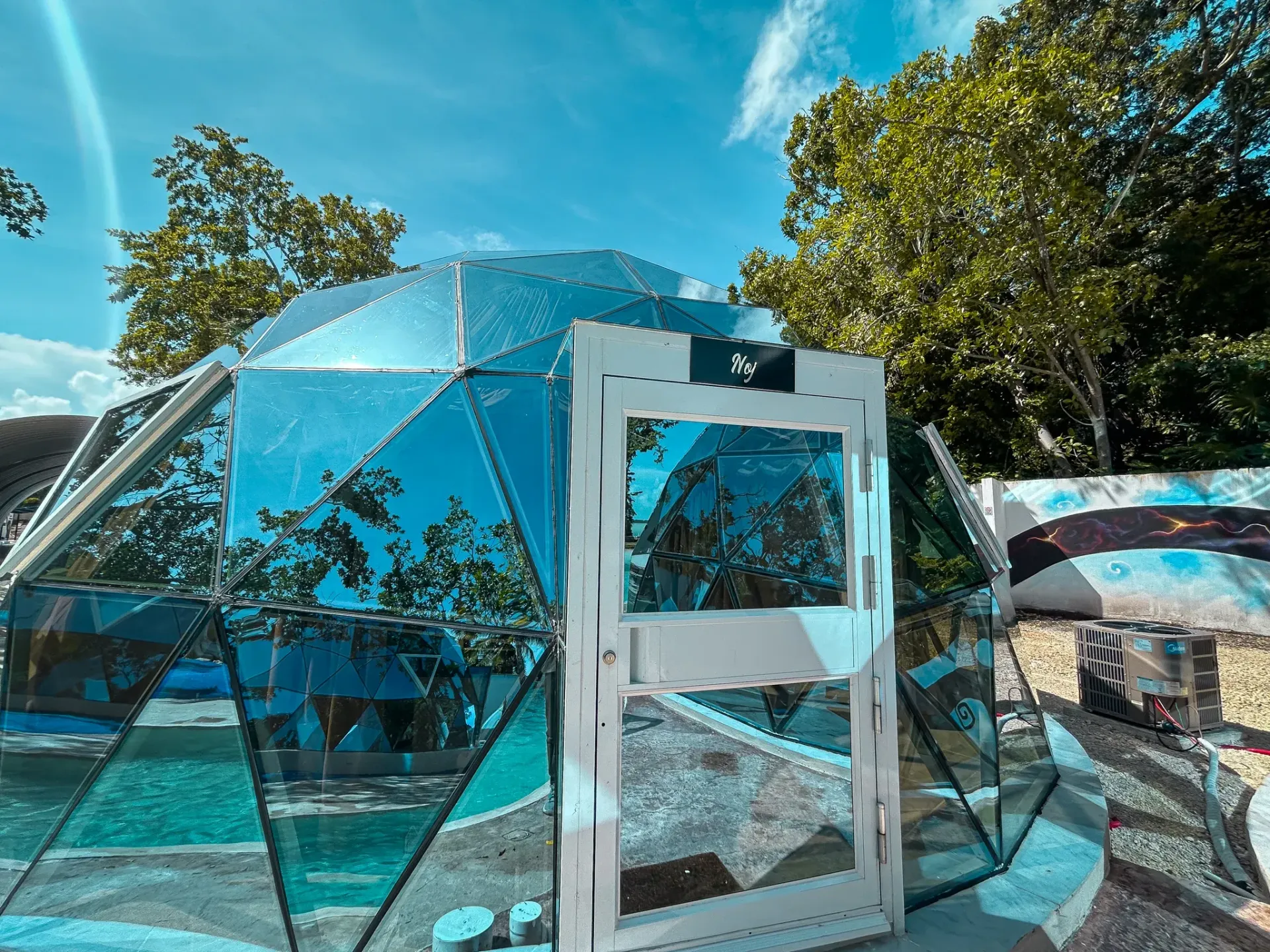 Cúpula geodésica con paneles de espejo y puerta blanca, con un fondo de árboles y cielo azul.