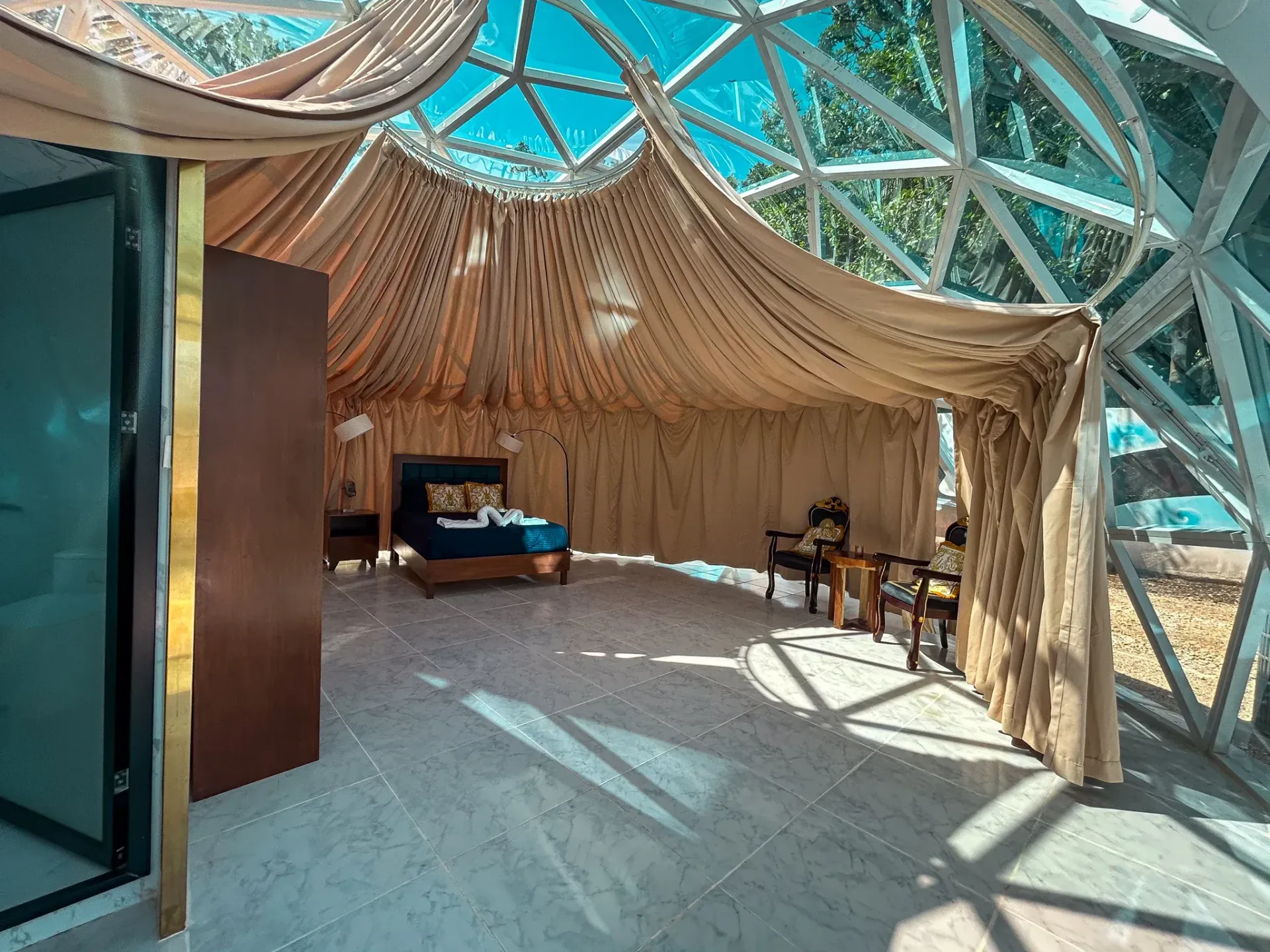Interior de cúpula geodésica con tela drapeada, cama y sillas; entra luz solar a raudales.