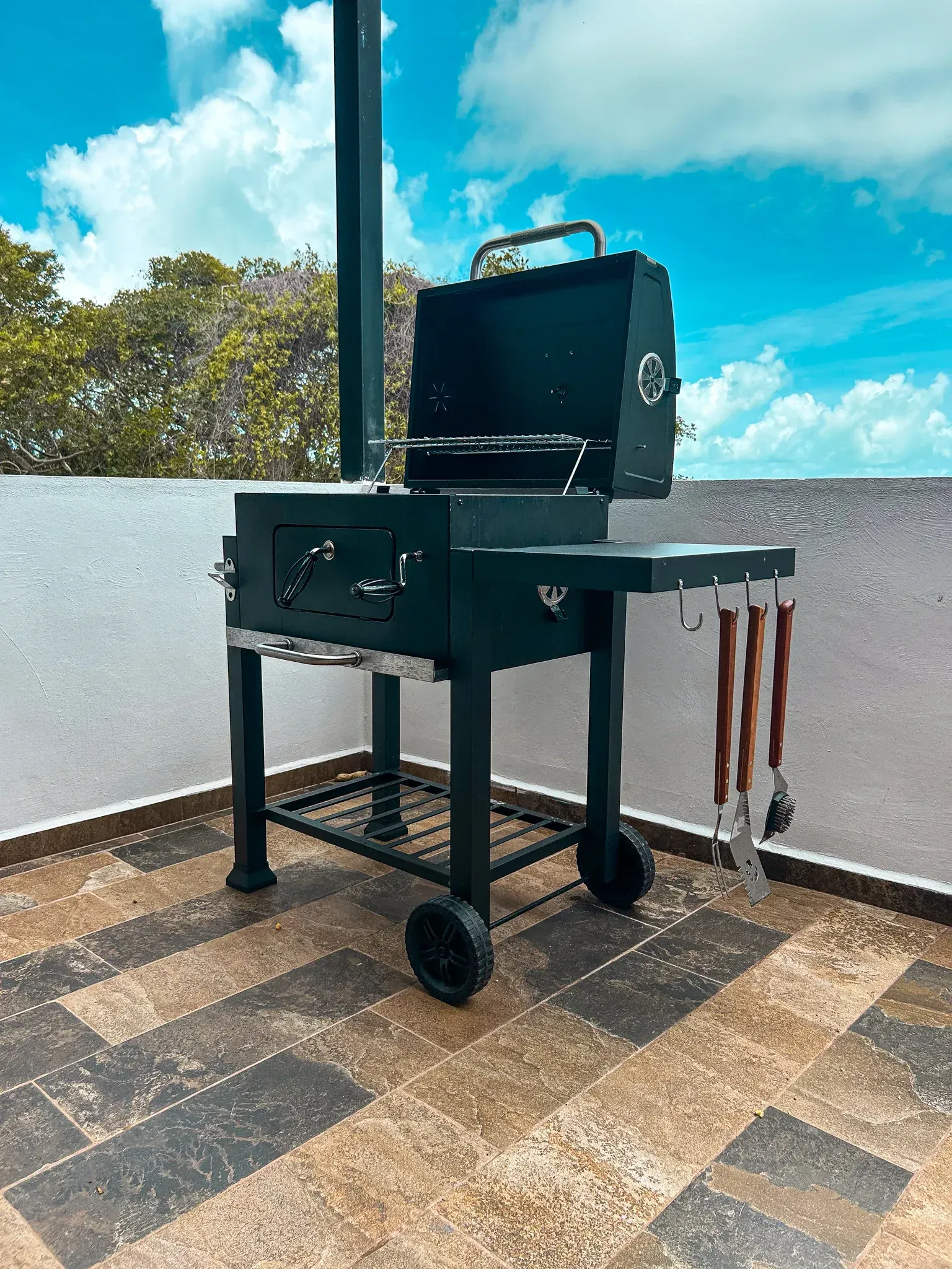 Parrilla de carbón negra sobre un patio de baldosas, con la tapa abierta y utensilios para asar colgando. 