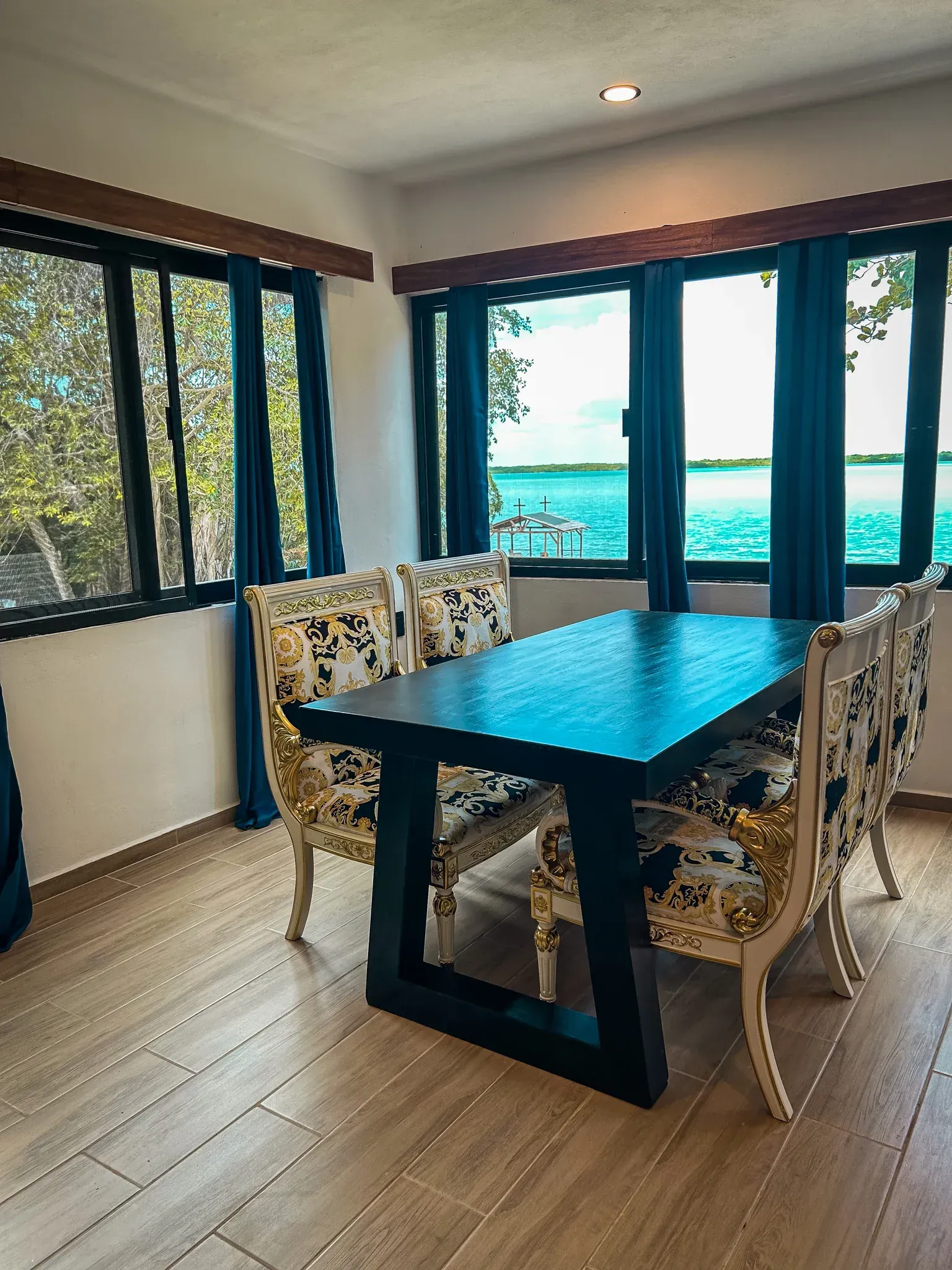 Comedor con mesa negra y sillas estampadas, ventanas con vistas al agua.