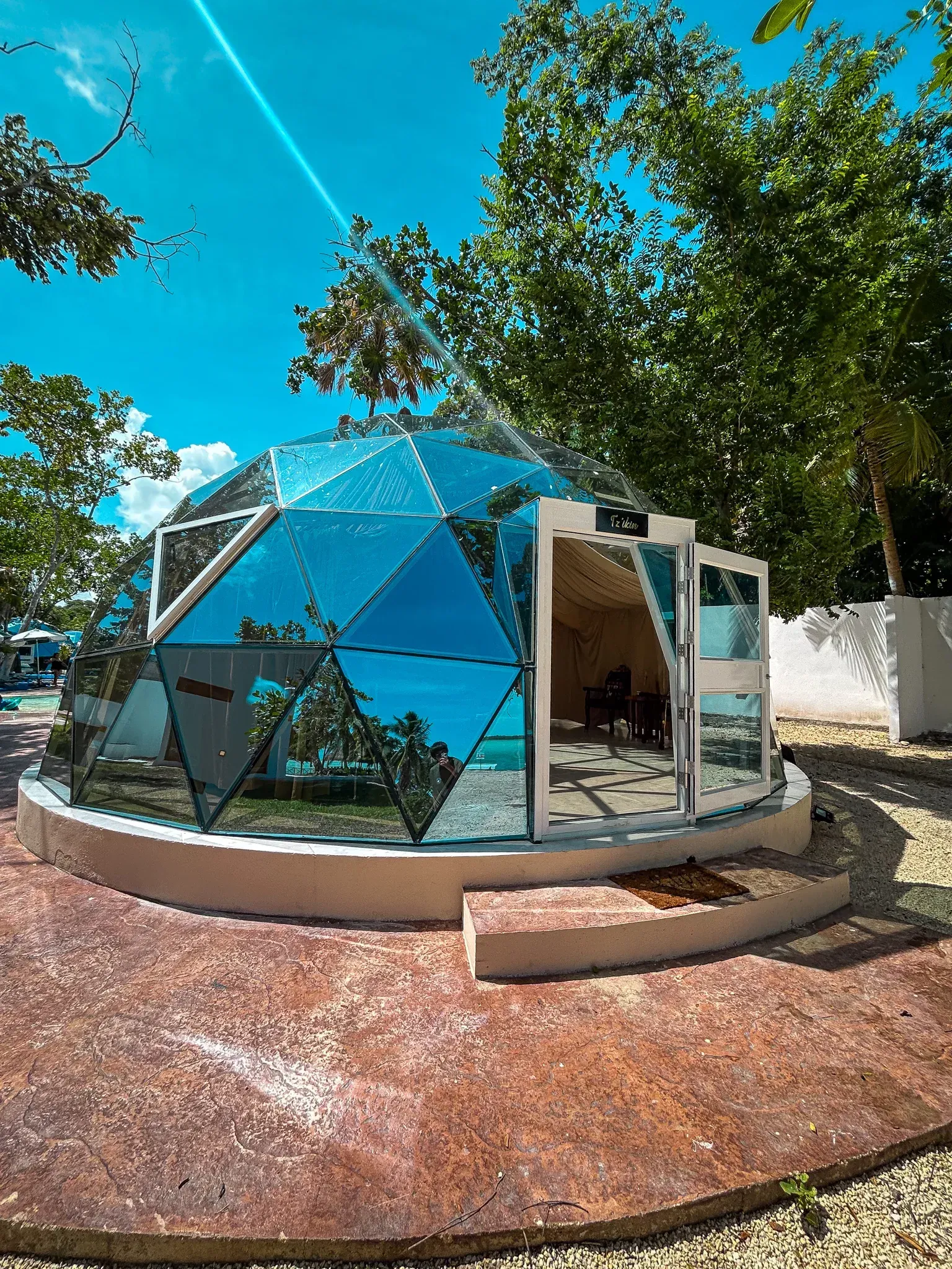 Glass dome structure with open doorway, reflecting blue sky and ocean. Brown and red flooring.