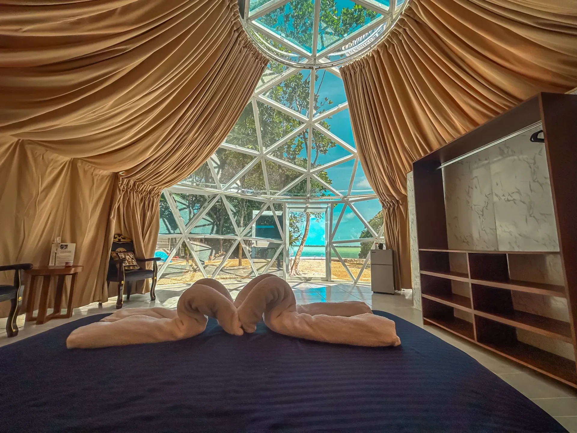 Interior of a dome-shaped luxury tent with swan towel art on a bed, looking out to a blue sky and trees.