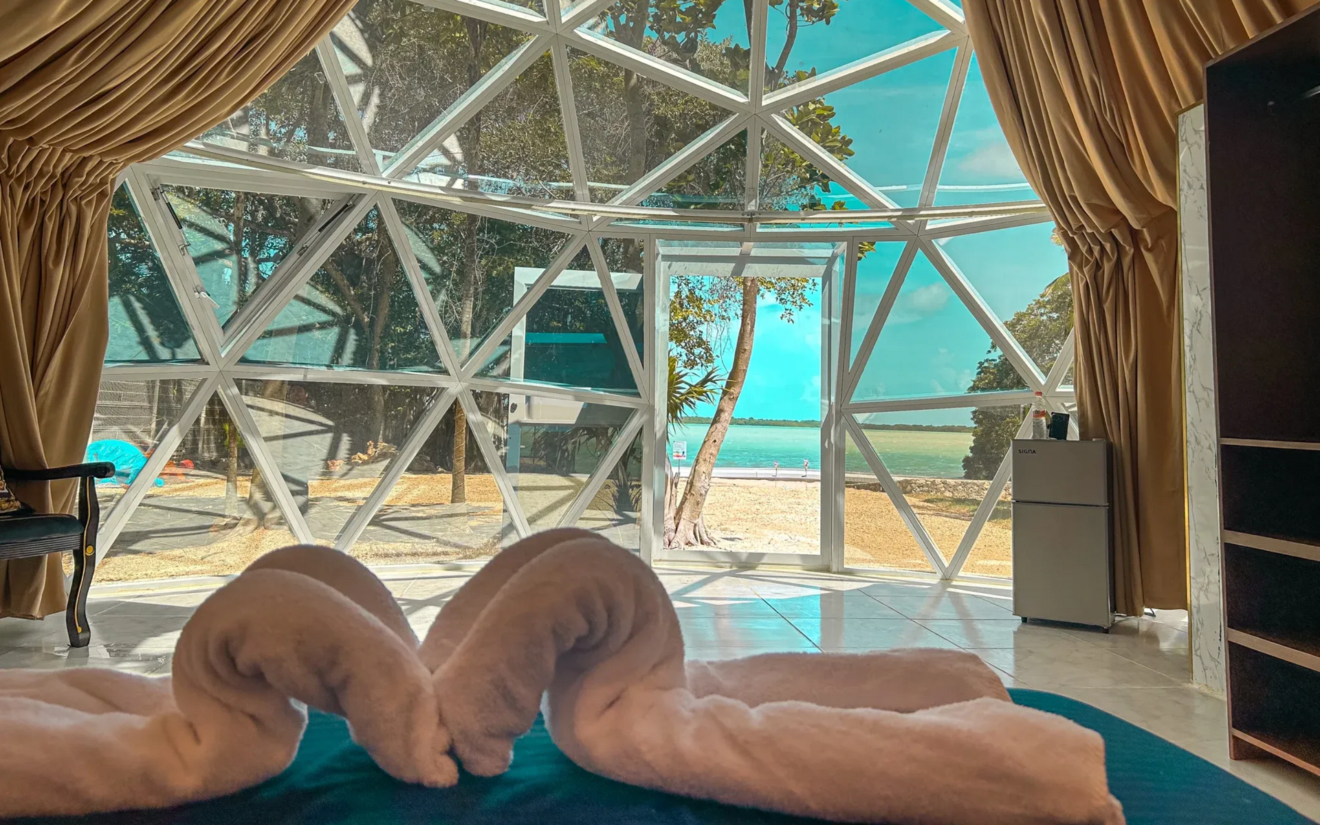 Interior de una cúpula geodésica con vistas a la playa. Dos toallas dobladas en forma de cisne sobre la cama.
