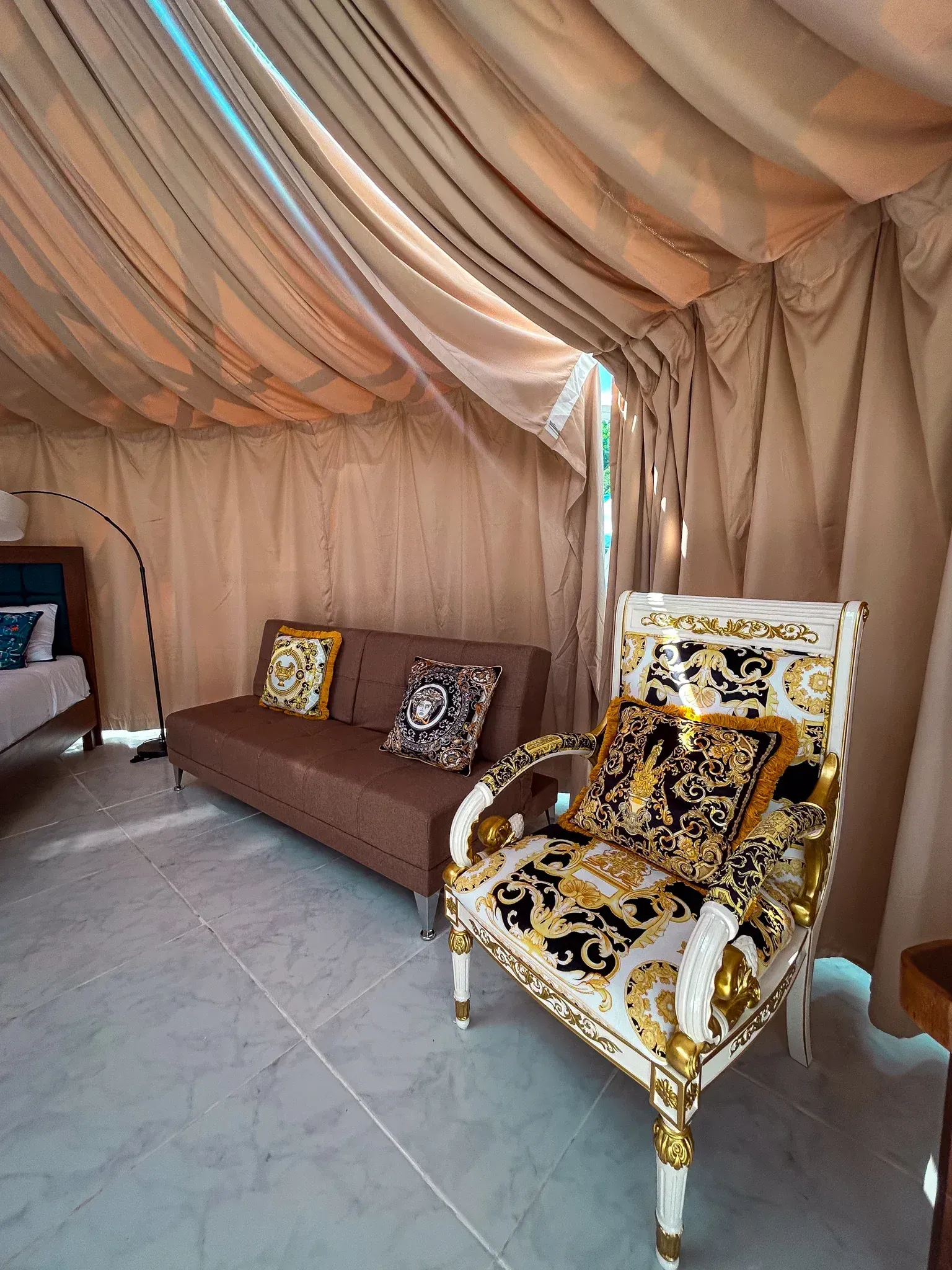 Interior de una tienda de campaña con sofá, silla ornamentada y cortinas. Combinación de colores beige claro y dorado.