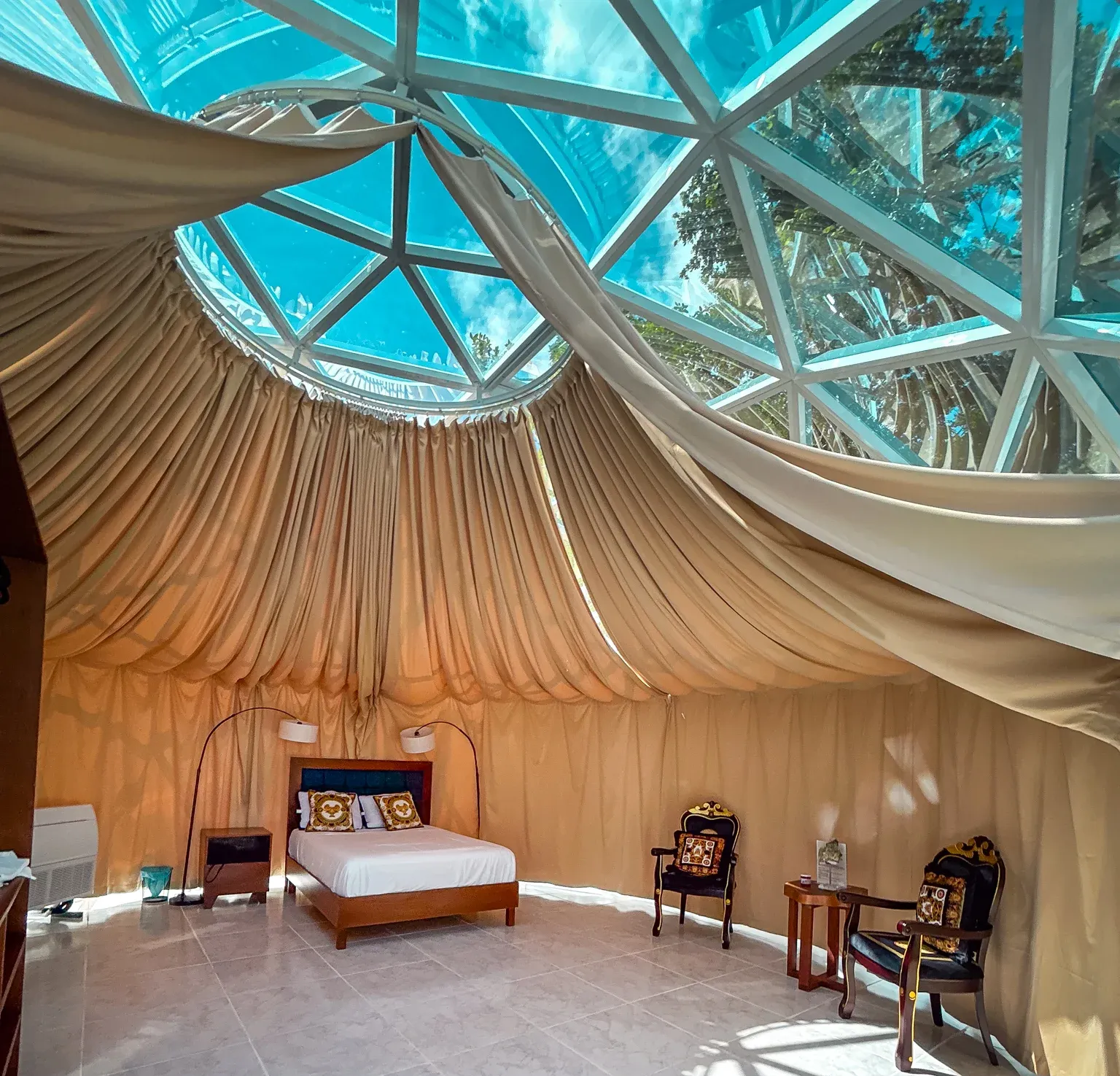 Un lujoso interior de yurta con techo abovedado transparente, cama, dos sillas ornamentadas y cortinas de tela color canela.