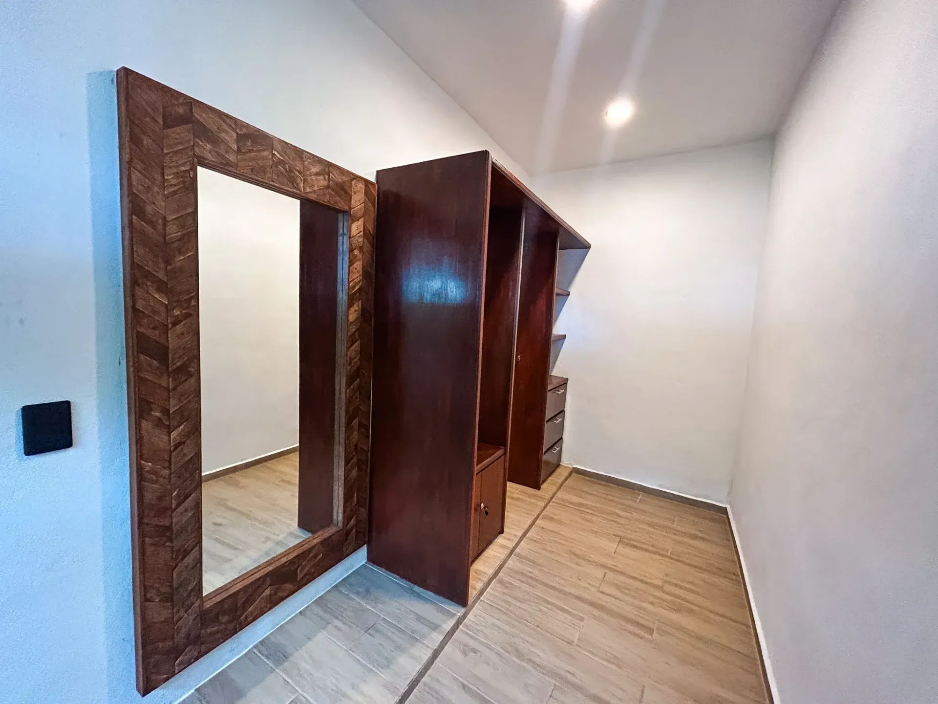 Vestidor con estantes de madera oscura y espejo con marco decorativo. Suelo de madera clara y paredes blancas.
