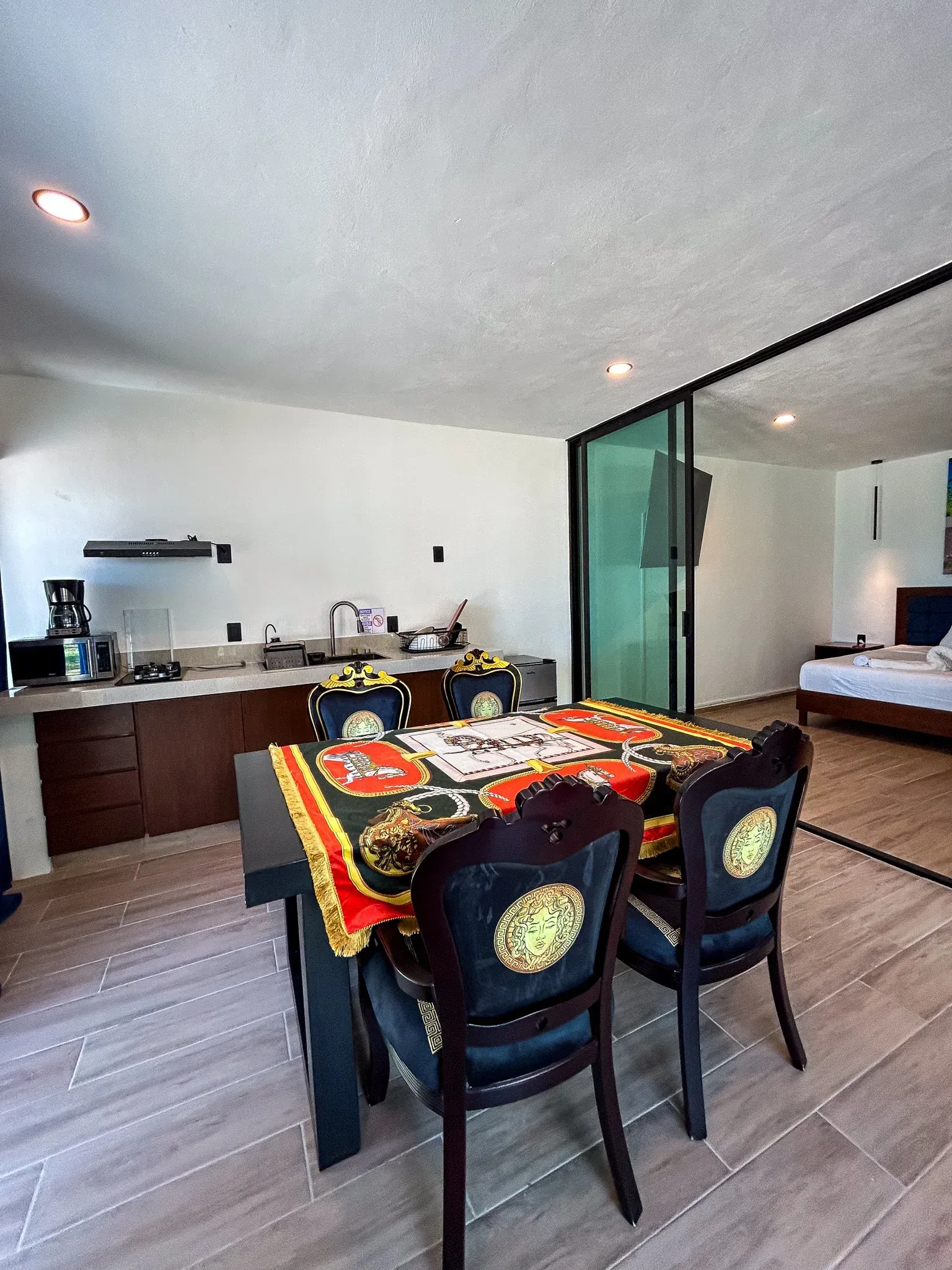 Comedor con mesa negra, sillas Versace, cocina y puerta de cristal que da al dormitorio.