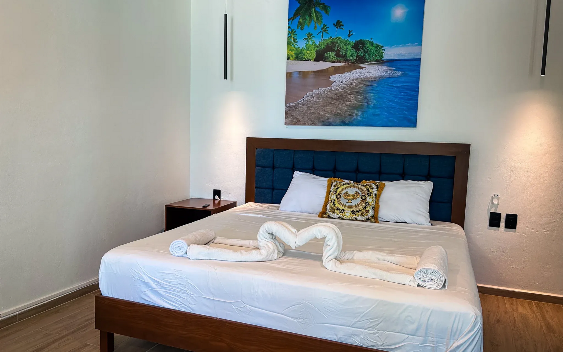 Dormitorio con cama, mesita de noche y cuadro con escena de playa. Sobre la cama hay dos toallas dobladas .