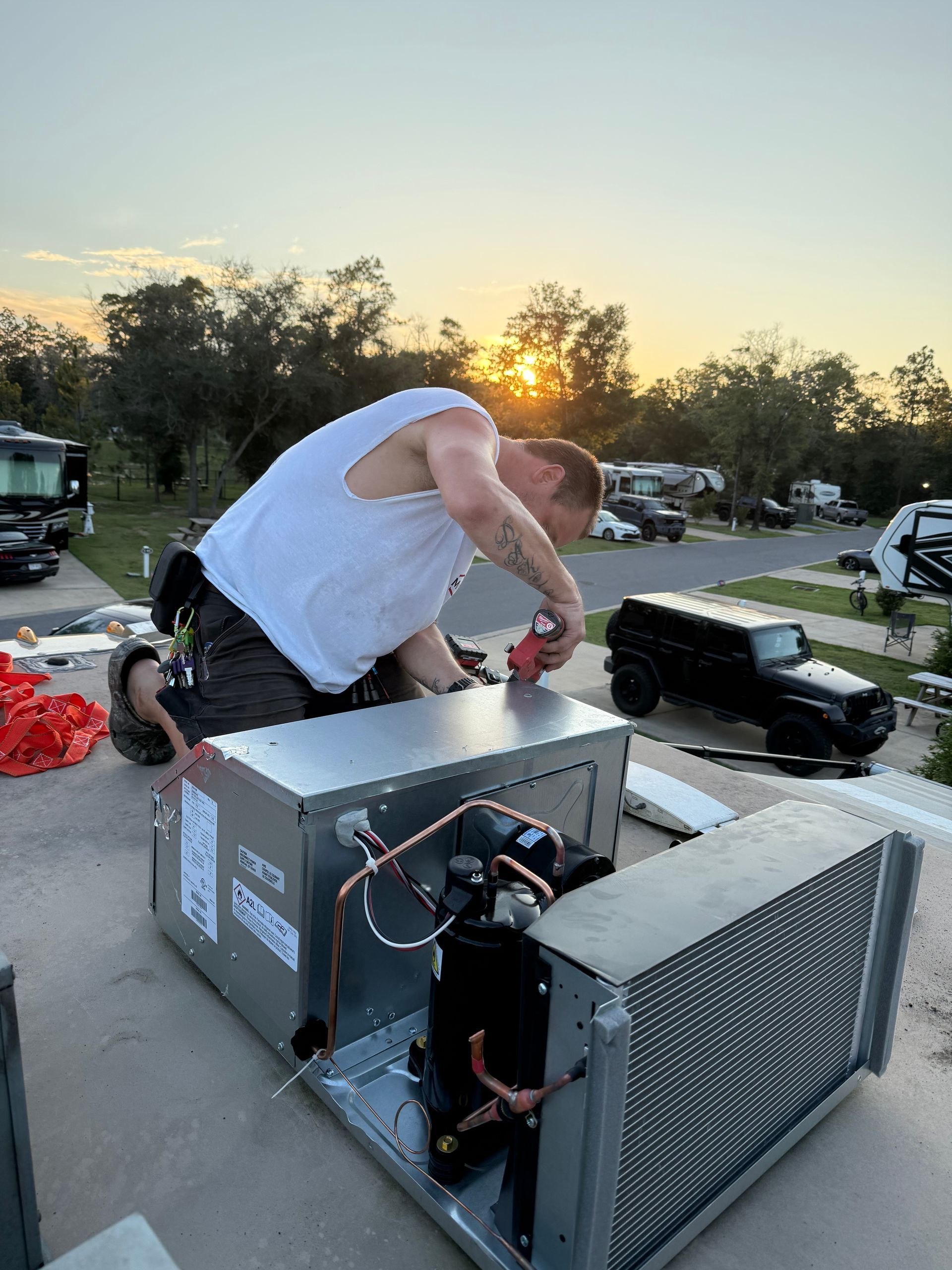 Person working on a metal machine outdoors at sunset beside trees and parked cars