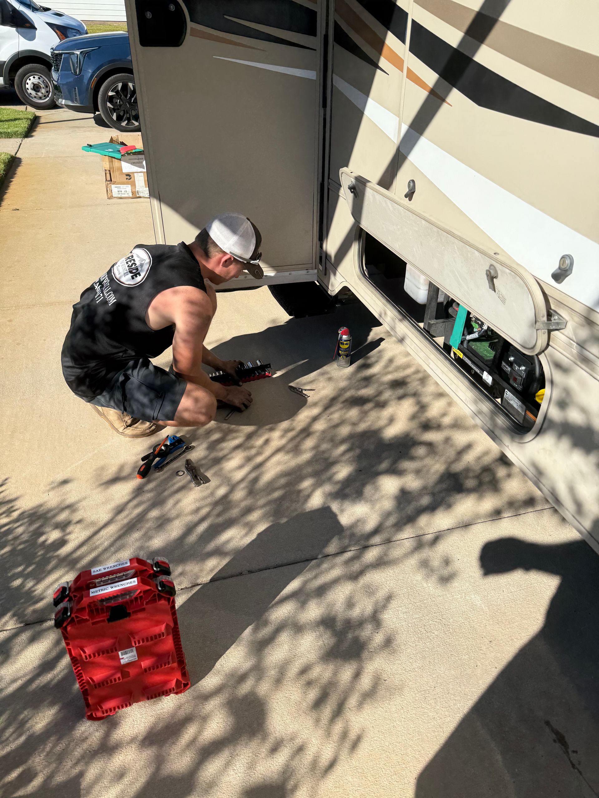 Person crouches beside an RV, working on equipment near the side panel in a sunny driveway.