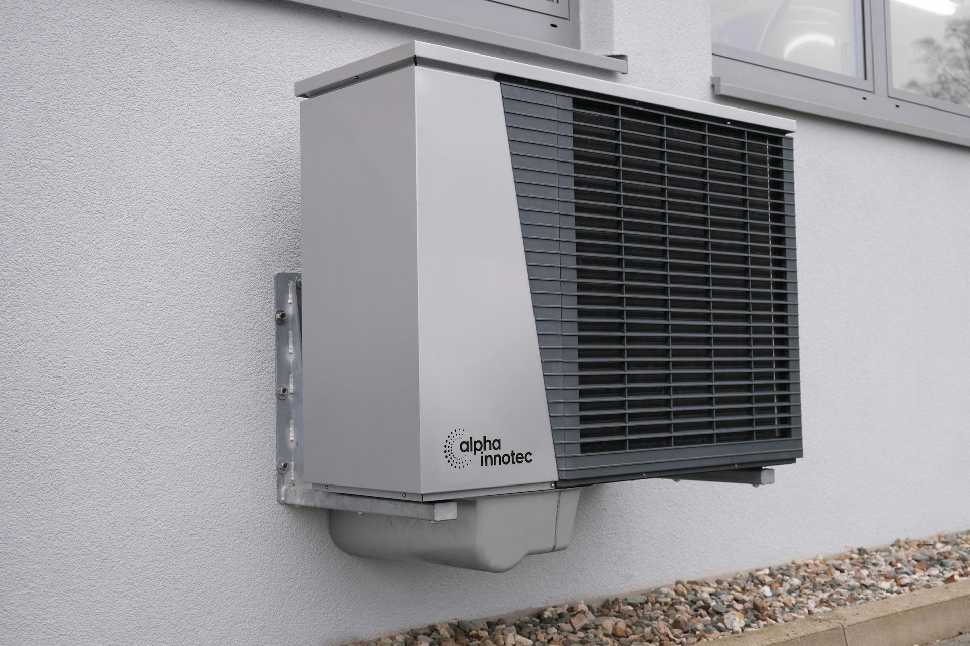 A grey, wall-mounted heat pump unit with dark grate. Mounted on white building next to a window.