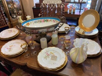Antique china and glassware displayed on a wooden table with floral plates, cups, and a serving bowl