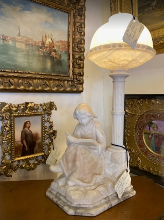 White statue on ornate shelf among framed paintings and a lamp in a decorative room