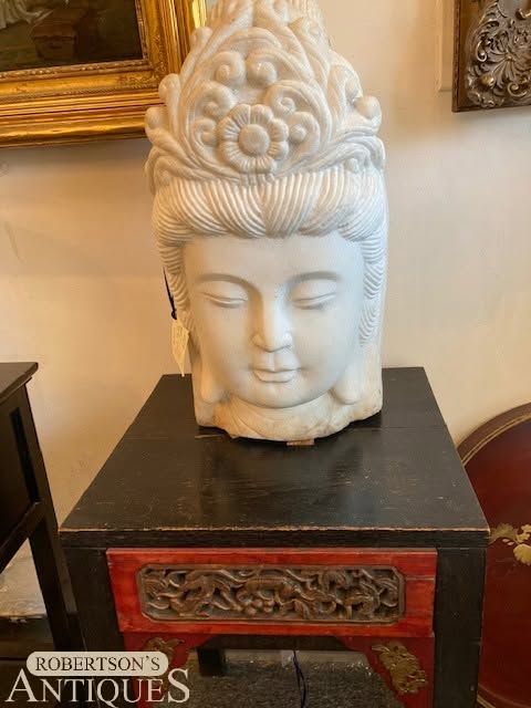 White Buddha head sculpture on a black pedestal with a red carved front panel in an antique shop