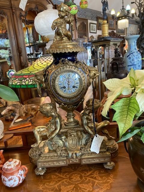 Ornate antique mantel clock with bronze cherubs and floral accents on a crowded antique shop display.