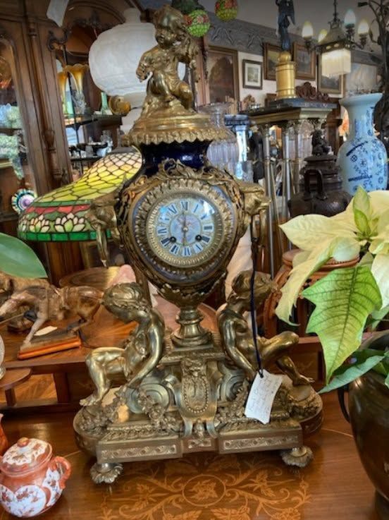 Ornate brass mantel clock with blue face and floral decorations on a carved wooden table in an antique shop
