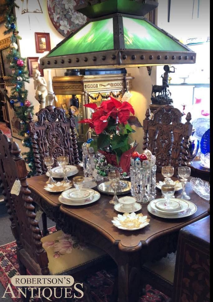 Antique dining table set with ornate chairs, green-shaded lamp, and red holiday centerpiece in a decorated room
