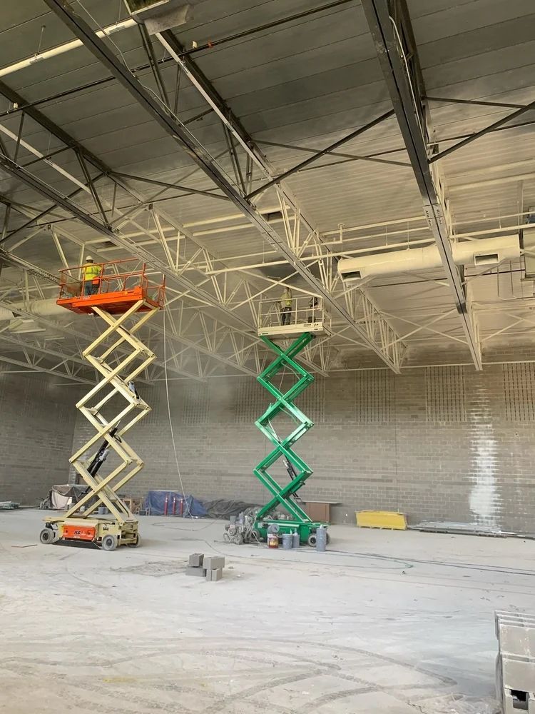 Two scissor lifts inside a large warehouse, one with a worker, painting the ceiling.