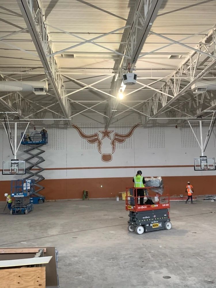 Gymnasium under construction with workers, scaffolding, and Longhorn logo on the wall.