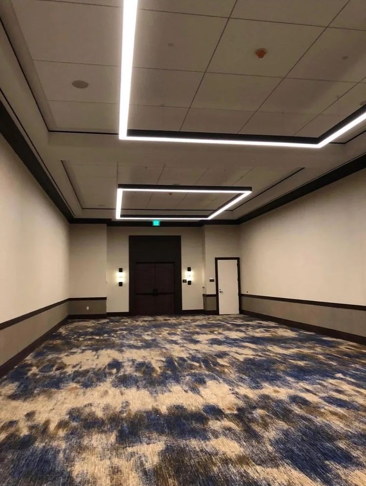 Empty event room with blue and brown patterned carpet, white walls, dark trim, and geometric ceiling lights.