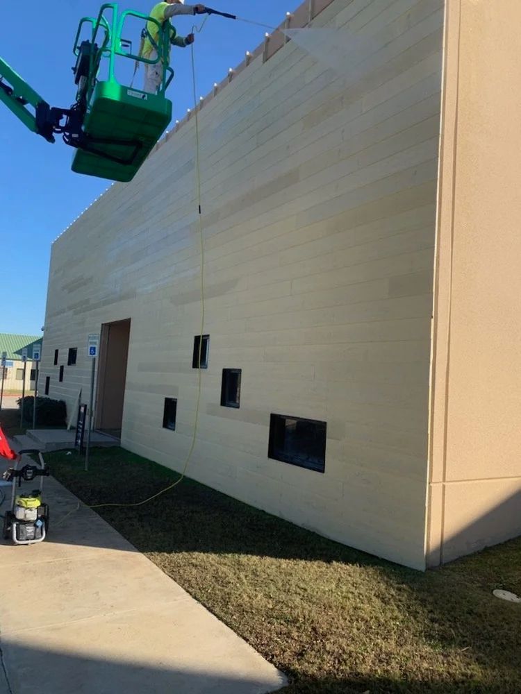 Person on a lift washes a light-colored building. Grass and sidewalk in front.