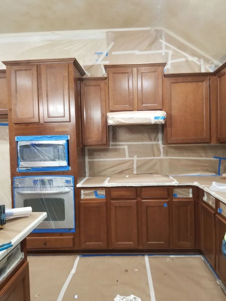 Kitchen cabinets in the process of being painted, with brown wood, covered appliances, and painter's tape.