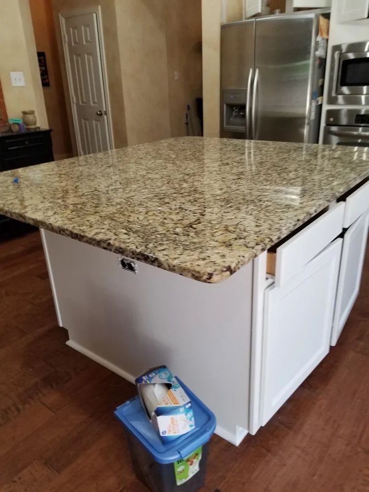 White kitchen island with granite countertop, wooden floor, and a stainless steel refrigerator.