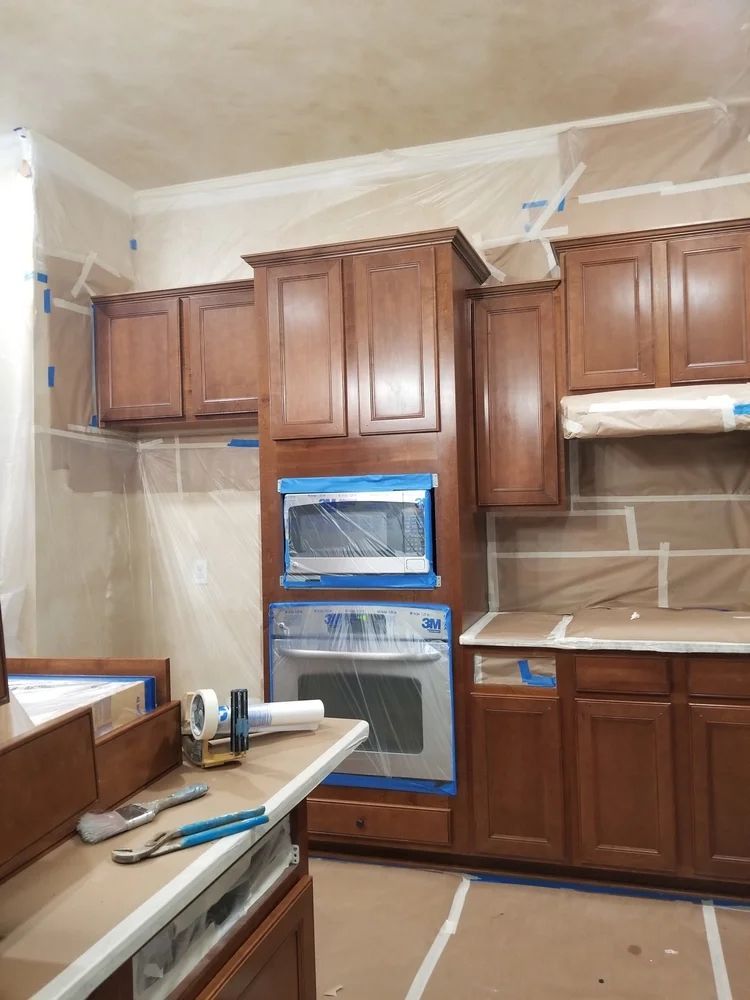 Kitchen cabinets and appliances being prepped for painting, covered in protective plastic and tape.