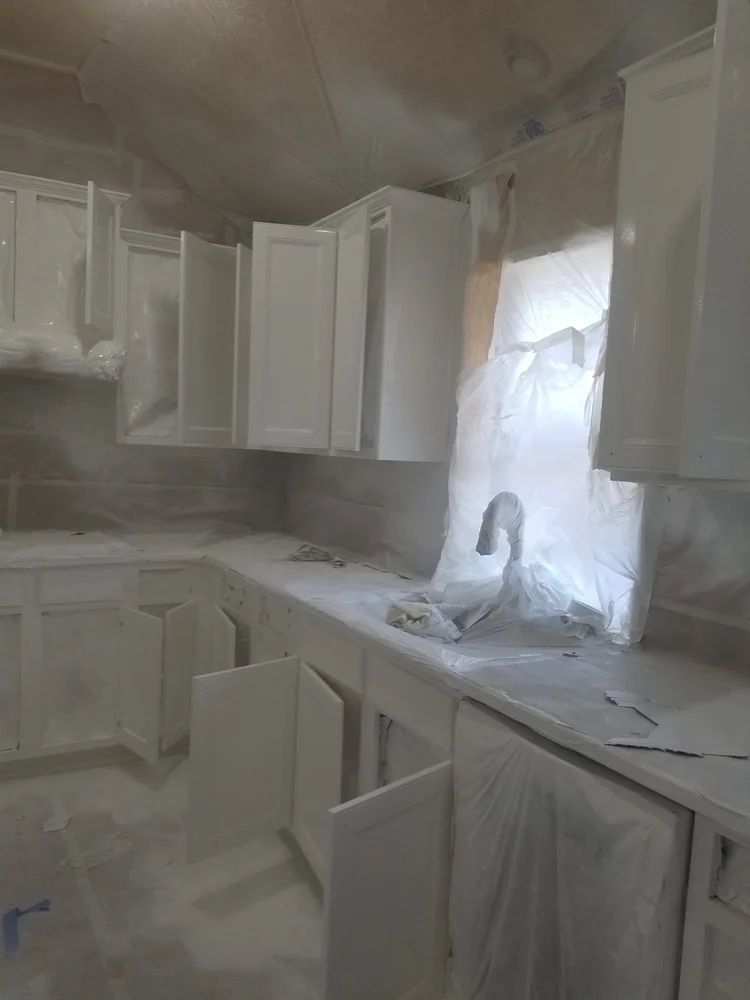 White kitchen cabinets and countertops covered in white paint spray during renovation.