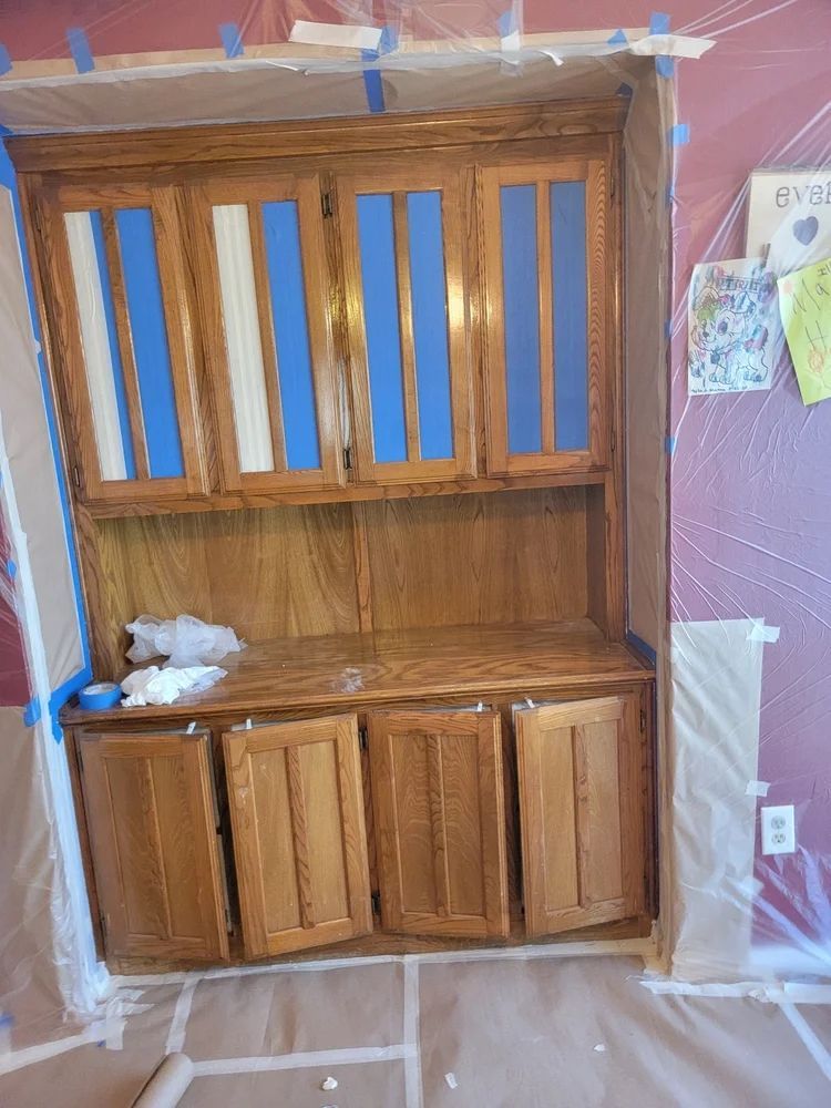 Wooden kitchen cabinet being painted; blue tape protects glass, surrounding area covered in plastic.