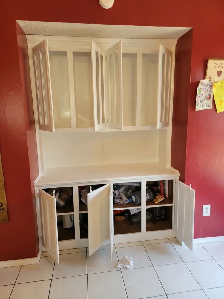 White built-in cabinet with open doors against a red wall.  Lower section stores items, upper is empty.