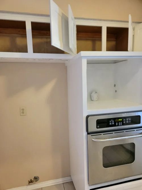 Kitchen cabinets with open doors, oven below, and a blank wall.