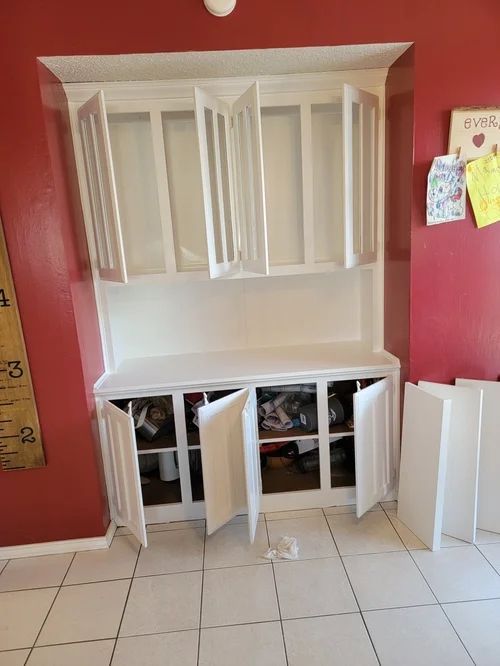 White built-in kitchen cabinet with open doors, against a red wall and tiled floor.