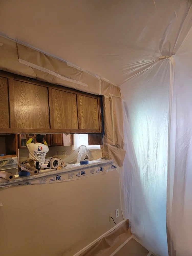 Room being remodeled, window with cabinets, plastic sheeting for containment. Beige wall, white paint buckets.