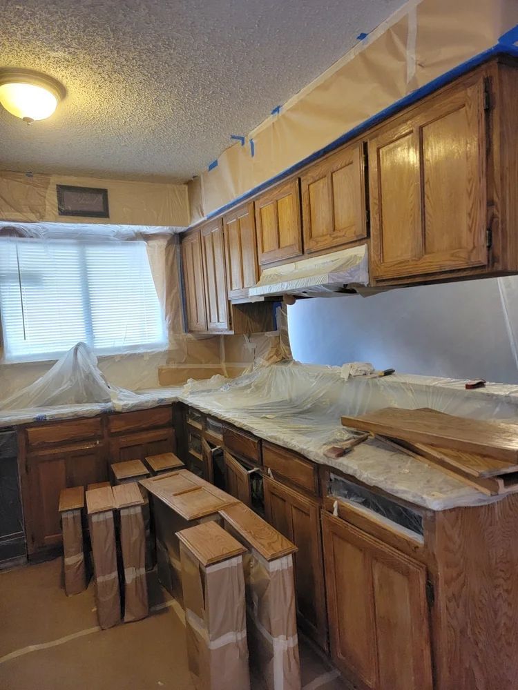 Kitchen cabinets being prepped for painting; plastic sheeting protects counters and walls, blue tape visible.