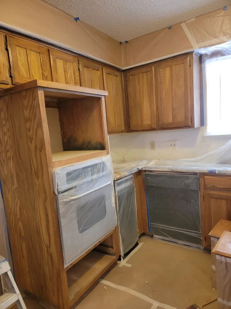 Kitchen cabinets, oven, and dishwasher covered in plastic during renovation, light brown wood.
