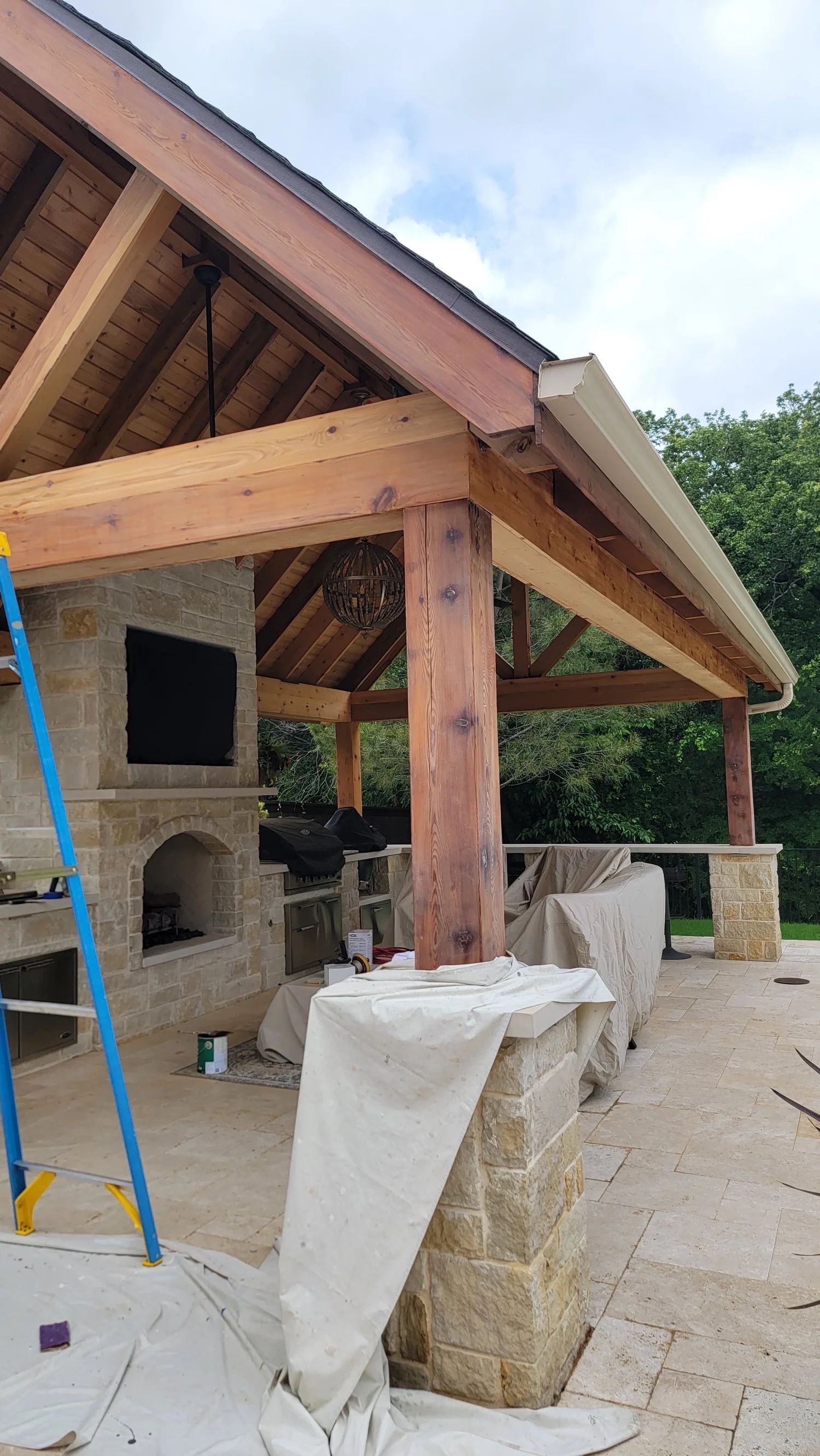 Outdoor pavilion under construction; wooden beams, stone pillars, fireplace, and ladder visible.